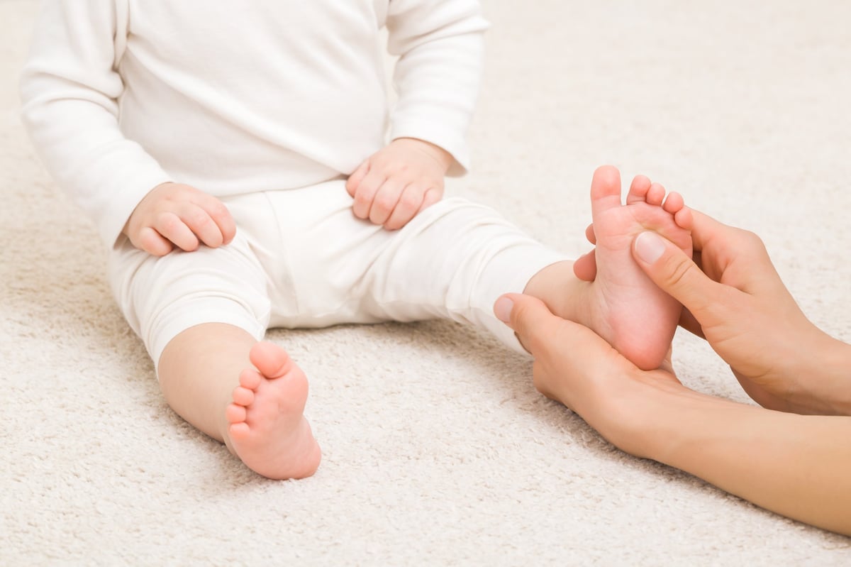 Hände eines Erwachsenen massieren sanft den Fuß eines Babys in weißer Kleidung auf hellem Teppich