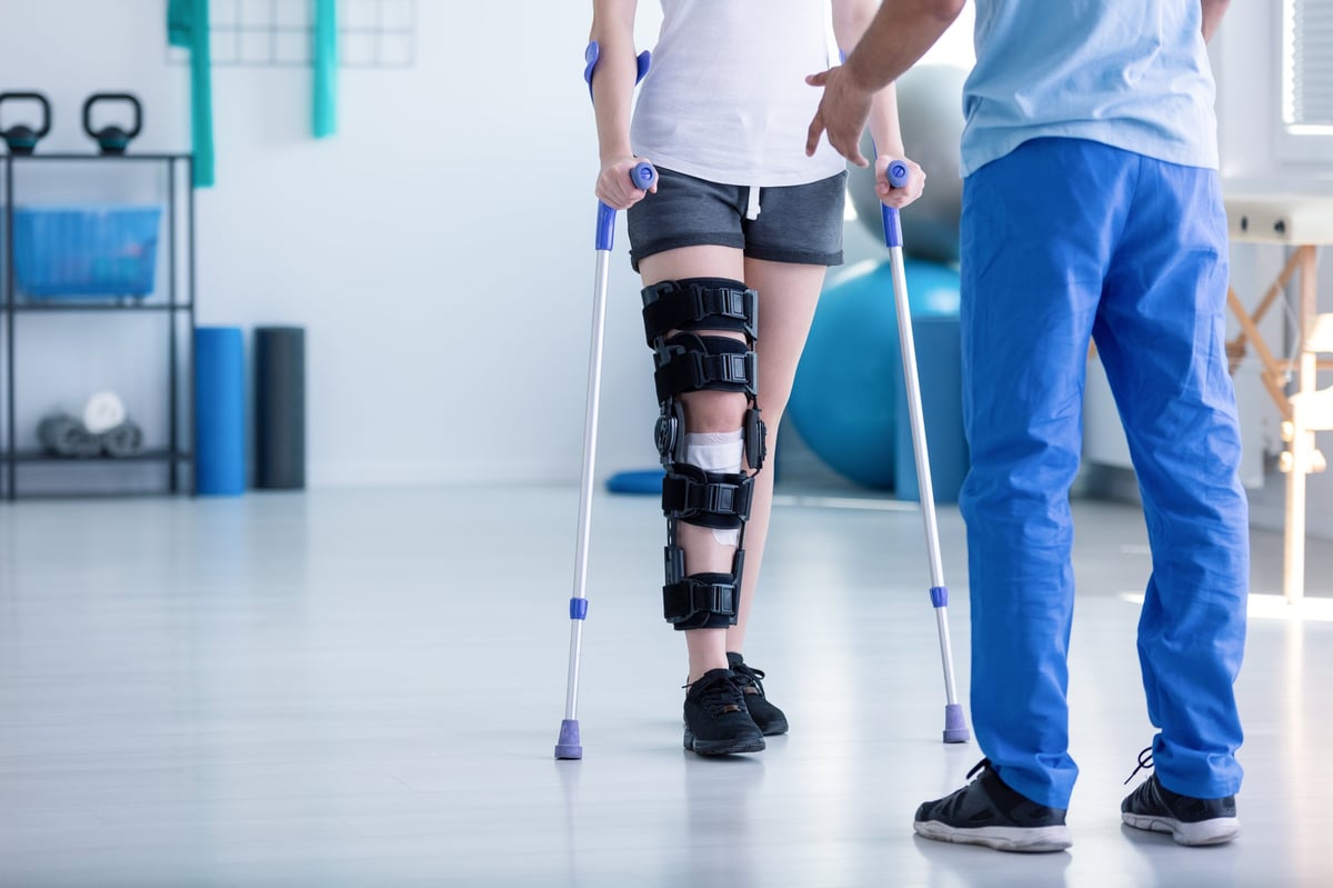 Patient mit Knieschiene an Krücken wird von einem Physiotherapeuten in einer Rehabilitationsklinik begleitet